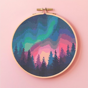 Könnte beinhalten: Ein Kreuzstich-Stickrahmen mit einer farbenfrohen Landschaft. Das Design zeigt eine Waldsilhouette unter einem lebendigen Nordlicht. Der Himmel wechselt von Blau zu Grün und Rosa. Der Holzrahmen wird von einem Messingverschluss gehalten.