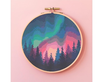 Puede incluir: Un bastidor de bordado de punto de cruz con un paisaje colorido. El diseño representa una silueta de bosque bajo una vibrante aurora boreal. El cielo transiciona de azul a verde y rosa. El bastidor de madera se sujeta con un cierre de latón.