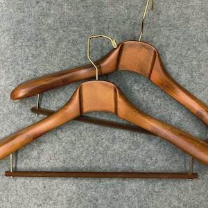 Op de afbeelding: Twee houten kledinghangers met een rijke, donkerbruine afwerking. De hangers hebben een gebogen ontwerp met een breed schoudergedeelte en een messingkleurige haak. Een houten stang is aan de onderkant bevestigd om broeken of rokken op te hangen.