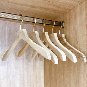 Pode incluir: Seis cabides de madeira com ganchos dourados pendurados em um armário. Os cabides são feitos de madeira marrom claro e têm um acabamento liso.