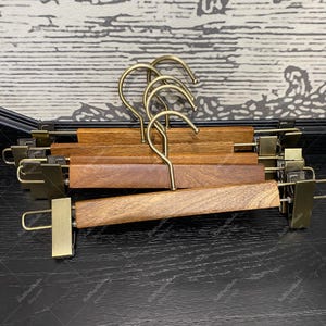 Op de afbeelding: Een set van vijf houten hangers met gouden metalen haken en clips. De hangers zijn gemaakt van donkerbruin hout en hebben een natuurlijke afwerking. De haken zijn gebogen en de clips zijn veerbeladen.