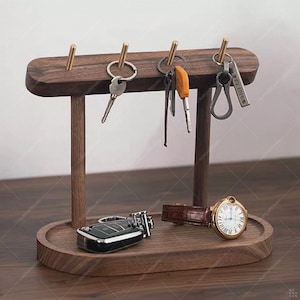 May include: A wooden key holder with a dark brown finish. The key holder has four brass hooks for keys and a lower tray. A car key fob and a gold-toned watch with a brown leather strap are on the tray.
