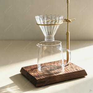 May include: A pour-over coffee maker featuring a clear glass dripper with a scalloped rim, a matching glass carafe with a handle, and a brass-toned metal stand. The entire assembly is mounted on a rectangular wooden base with a textured surface.