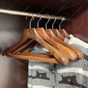 Puede incluir: Conjunto de seis perchas de madera con un acabado marrón natural y ganchos dorados. Las perchas están apiladas una encima de la otra y están colgadas en un armario.