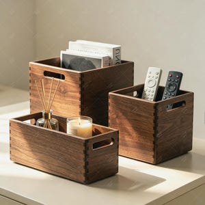 May include: Three rectangular dark brown wooden storage boxes with finger-jointed corners and cutout handles. The largest box holds books, the medium box contains two remote controls, and the smallest box holds a glass candle and reed diffuser.