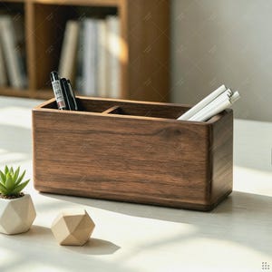 Puede incluir: Un organizador de escritorio rectangular de madera marrón oscuro con dos compartimentos internos. El lado izquierdo contiene dos bolígrafos negros, mientras que el lado derecho contiene tres bolígrafos blancos. La madera muestra vetas visibles y bordes redondeados.