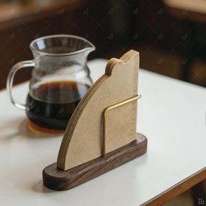 Könnte beinhalten: Ein Stapel beigefarbener Kaffeefilter aus Papier, die von einem Messingdrahtrahmen auf einem dunkelbraunen Holzsockel aufrecht gehalten werden. Eine klare Glaskaraffe mit dunkelbraunem Kaffee steht hinter dem Filterhalter auf einer weißen Fläche.
