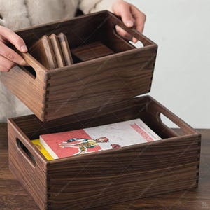 Op de afbeelding: Twee houten opbergdozen met handvatten, waarvan één opgetild om de inhoud te onthullen. De bovenste doos bevat kleine houten blokken, terwijl de onderste doos een boek met een rode en witte kaft bevat. De dozen zijn diep bruin van kleur.