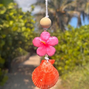 May include: A hanging ornament with a beach theme, featuring a bright pink hibiscus flower, a natural wooden bead, and a vibrant orange seashell. The ornament is suspended by a white string, creating a tropical and summery aesthetic.