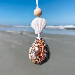 Pode incluir: Um móbile de conchas com uma concha branca, uma conta de madeira e uma concha marrom e branca penduradas em um cordão branco.