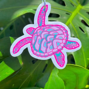 Könnte beinhalten: Ein rosa-türkisfarbener Meeresschildkröten-Lufterfrischer mit weißem Hintergrund. Die Schildkröte ist stilisiert mit einem detaillierten Panzer und Flossen.