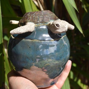 May include: A round ceramic jar with a glossy blue glaze and a textured brown lid. A sculpted clay turtle with a light-colored head and limbs is attached to the top of the lid, appearing to emerge from the center of the blue ceramic vessel.