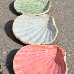 May include: Three ceramic shell-shaped dishes in a row. The top dish is light green, the middle is light blue, and the bottom is pink. Each dish has a textured, ridged surface and a rough, unglazed rim. They are displayed on a sandy surface.