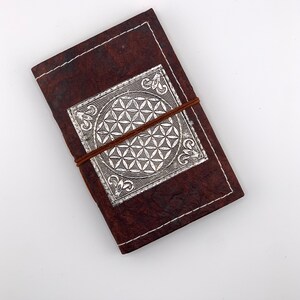 May include: A brown leather journal with a silver embossed flower of life design. The journal is closed and has a brown leather strap.