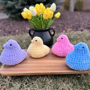 Könnte beinhalten: Vier Plüsch-Küken-Spielzeuge in Lavendel, Gelb, Rosa und Blau sitzen auf einem Holztablett. Im Hintergrund steht ein schwarzer Topf mit gelben und weißen Tulpen. Die Spielzeuge sind handgefertigt und haben eine weiche Textur.