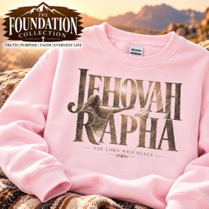 May include: A light pink crewneck sweatshirt featuring a graphic print with the text JEHOVAH RAPHA and THE LORD WHO HEALS. The design includes an illustration of a person reaching out, integrated into the bold, dark lettering.