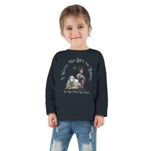 May include: Navy blue long-sleeve shirt with a graphic of a young boy, a sheep, and the text "I HAVE NO GIFT TO BRING, PA RUM PUM PUM PUM". The design includes a star. The shirt is worn by a child.