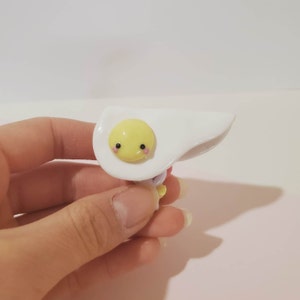 May include: A white and yellow clay figurine of a fried egg with a cute cartoon face. The egg has a white yolk and a pink blush.