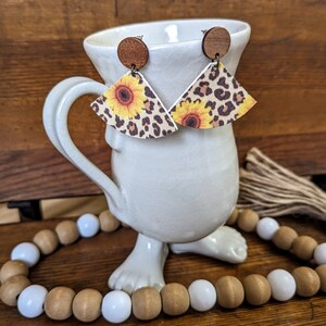 May include: A pair of dangle earrings with a brown wooden top and a fan-shaped bottom. The bottom is made of faux leather with a leopard print and a sunflower design.