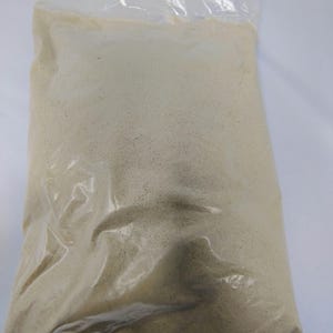 May include: A clear plastic bag filled with a light tan, granular substance. The bag is sealed at the top, and the contents appear to be a fine powder or small granules. The bag is slightly translucent, allowing the contents to be visible.