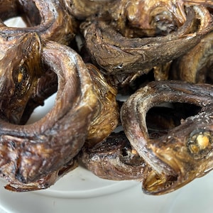 May include: Close-up of a pile of dried, smoked fish. The fish are a deep brown color with a slightly glossy texture. The fish are arranged in a circular shape, with the heads and tails curled inwards. The fish are on a white plate.