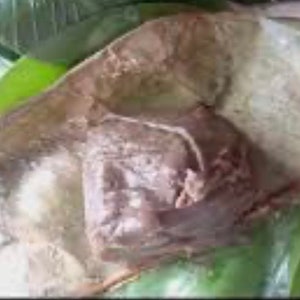 May include: A close-up of a large, light brown, irregularly shaped food item wrapped in a white, translucent material. The food is sitting on a bed of green banana leaves.