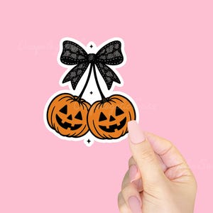 May include: A sticker featuring two orange jack-o'-lanterns with black carved faces, connected by black stems to a black lace bow. The sticker is on a pink background. The design has a Halloween theme.