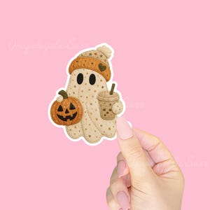 May include: A whimsical sticker featuring a friendly ghost wearing a knit hat and holding a jack-o'-lantern and a boba tea. The ghost is beige with star patterns. The pumpkin is orange, and the boba tea is brown. The background is pink.