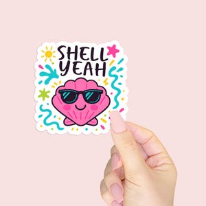 May include: A colorful sticker featuring a pink seashell wearing sunglasses with the words "SHELL YEAH". The design includes a sun, stars, and water splashes. The sticker is held against a light pink background.
