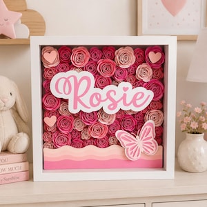 Puede incluir: Un marco cuadrado blanco tipo caja con un arreglo denso de rosas de papel en tonos rosa y rojo. El nombre Rosie está escrito en letra cursiva blanca sobre un recorte en forma de nube, junto a una mariposa de papel y corazones pequeños.