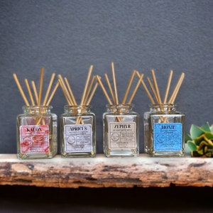 May include: Four glass reed diffusers with wooden reeds, each with a different label: KALON, APRICUS, ZEPHYR, and MOXIE. The diffusers sit on a wooden shelf against a dark gray background. Each label has text and a small illustration.