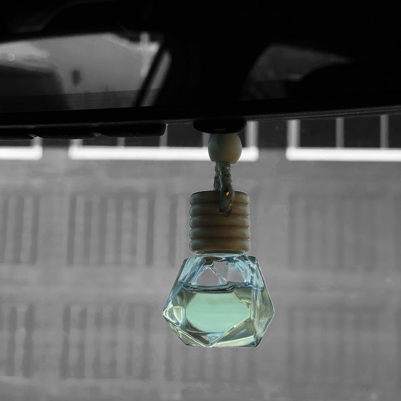 May include: A clear glass car air freshener with a wooden cap and a string hanging from it. The air freshener is hanging from the rearview mirror of a car.