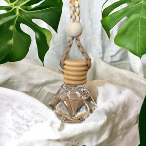 May include: A clear glass diamond-shaped bottle with a wooden cap and a beige and gold cord. The bottle is hanging from a white surface with green leaves in the background.