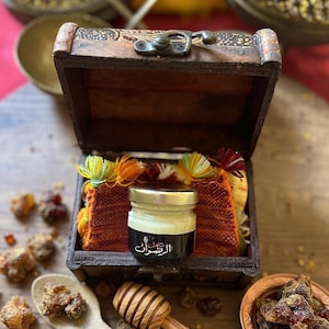 Puede incluir: Un pequeño frasco de vidrio con tapa plateada y etiqueta negra con texto árabe en blanco, colocado dentro de un cofre de madera oscura. El cofre contiene una tela naranja texturizada con borlas coloridas. Un cucharón de miel descansa cerca.
