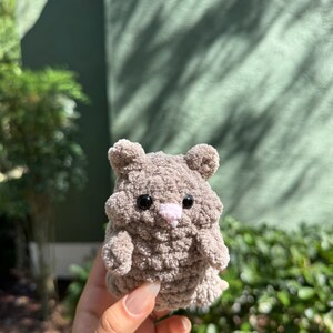 May include: A small, gray crocheted stuffed animal resembling a hamster. It has black eyes, a pink nose, and a fluffy texture.