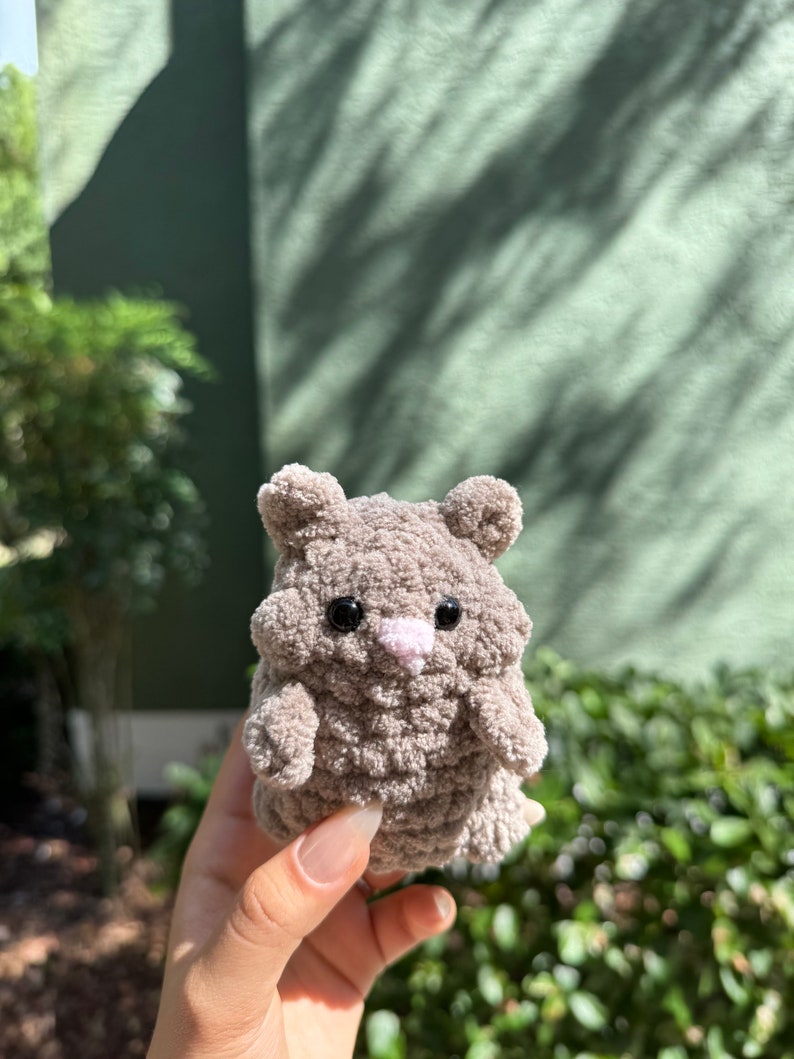 May include: A small, gray crocheted stuffed animal resembling a hamster. It has black eyes, a pink nose, and a fluffy texture.