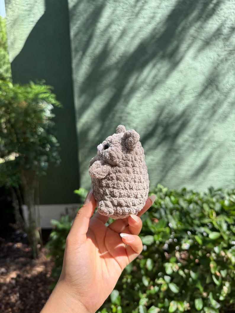 May include: A small, gray crocheted stuffed animal shaped like a bear. The bear is being held in a person's hand.