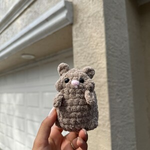 May include: A small, brown crocheted hamster toy with pink nose and eyes. The toy is being held in a person's hand.