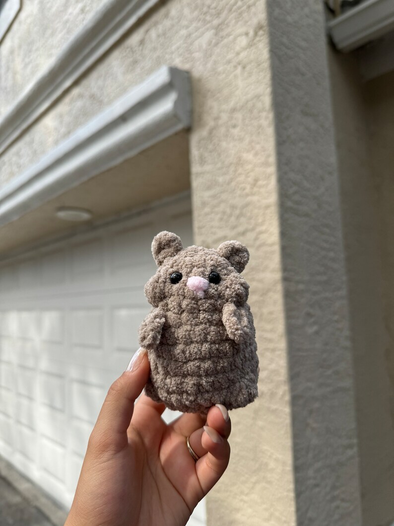 May include: A small, brown crocheted hamster toy with pink nose and eyes. The toy is being held in a person's hand.