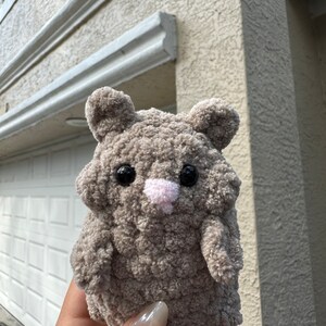 May include: A small, gray, crocheted stuffed animal shaped like a hamster. It has black eyes, a pink nose, and a fluffy texture.