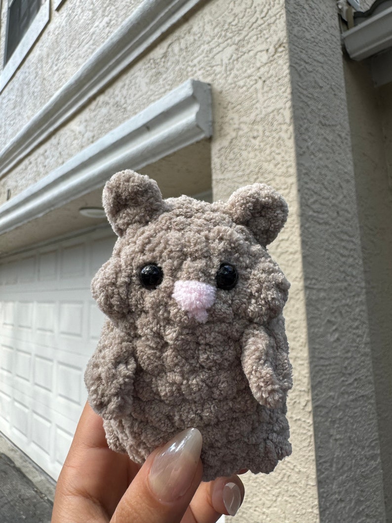May include: A small, gray, crocheted stuffed animal shaped like a hamster. It has black eyes, a pink nose, and a fluffy texture.