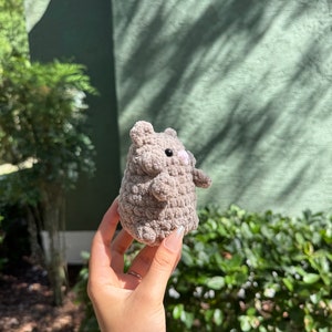 May include: A small, gray crocheted stuffed animal with a pink nose and black eyes. The animal is being held in a person's hand.