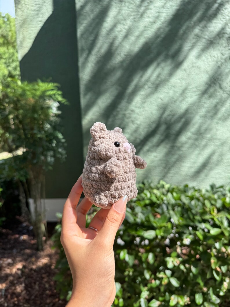 May include: A small, gray crocheted stuffed animal with a pink nose and black eyes. The animal is being held in a person's hand.