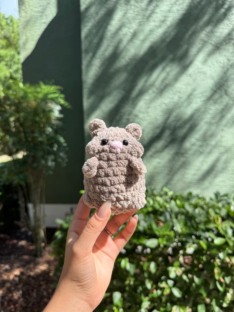 May include: A small, crocheted hamster toy in a light brown color. The toy has black eyes and a pink nose.