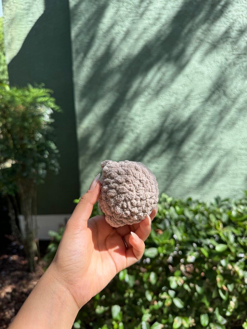 May include: A close-up of a gray, textured, round crocheted object. The object is being held in a person's hand.