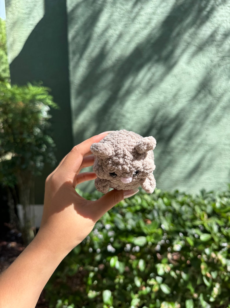 May include: A small, gray, plush toy shaped like a bear with a pink nose and black eyes. The toy is being held in a person's hand.