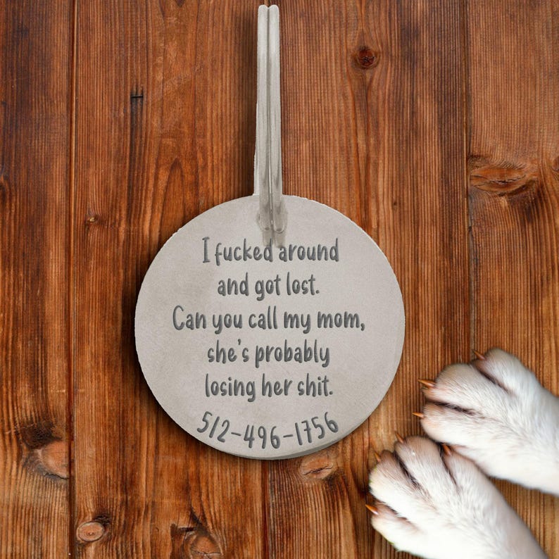 Silver engraved pet tag displayed on wooden background with dog paws in frame. Round ID tag reads “I fcked around and got lost. Can you call my mom, she’s probably losing her sht.” with phone number engraved below. Funny yet practical pet accessory for dogs who wander, offering safety and style. Durable metal design with custom etching, ideal gift for dog moms and pet lovers wanting humor and function.