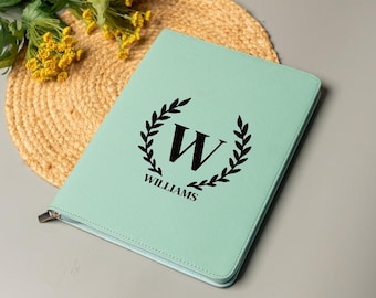 May include: A light green leather portfolio with a black wreath design, the letter "W", and the name "WILLIAMS" printed on the front. The portfolio has a zipper closure and is lying on a woven mat.