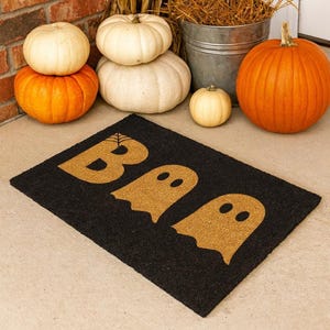 May include: Black rectangular doormat with the word "BOO" in gold letters and ghost shapes. The mat is surrounded by pumpkins and gourds in shades of orange and white, creating a festive autumn display.