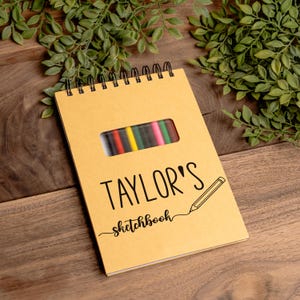 A personalized spiral-bound sketchbook engraved with the name “Taylor’s Sketchbook” on a tan cover, displayed on a wooden surface with green leaves for decoration. The cover features a pencil graphic and a cut-out window revealing 12 colorful pencils inside, stored in an attached pouch. This custom sketchbook makes a thoughtful and practical gift for kids, teens, students, or artists, combining creativity, organization, and personal style in one unique art journal.